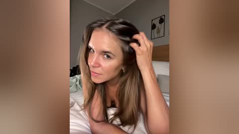 A video of a nude woman with long, straight, light brown hair, lying on a bed in a dimly lit bedroom. Her breasts are partially visible.