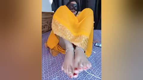 Video of a woman in a bright yellow sari with intricate lace trim, seated on a purple patterned bedspread, wearing anklets and toe rings, with her feet prominently in the foreground.