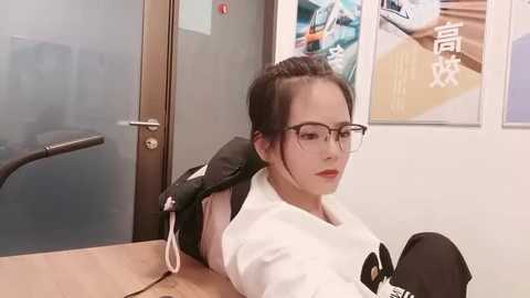 Video of a young Asian woman with glasses, fair skin, and dark hair tied back, wearing a white martial arts uniform, leaning against a wooden desk in an office.