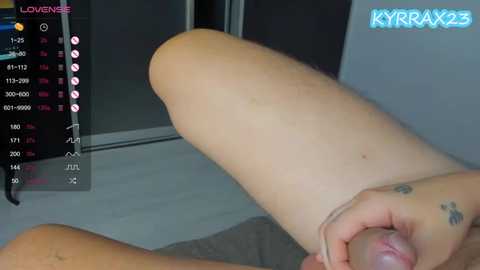 Video of a nude person with light skin, a tattoo on their left hand, and a visible erect penis, lying on a bed with a gray sheet. Background includes a TV displaying the KYRAZX3 profile and a digital clock.
