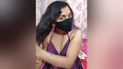 Media: A video of a woman with long black hair, wearing a purple lace bralette, black face mask, and silver bracelets, holding her hair while seated on a bed with a pink floral sheet.