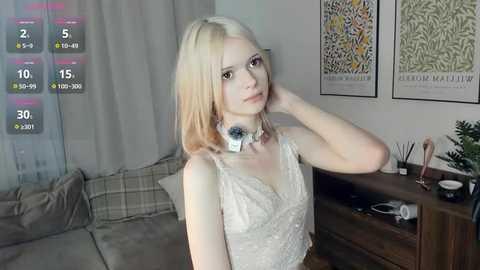 Video of a young, fair-skinned woman with shoulder-length blonde hair, wearing a white lace dress, standing in a cozy living room with plaid pillows, artwork, and a wooden console.
