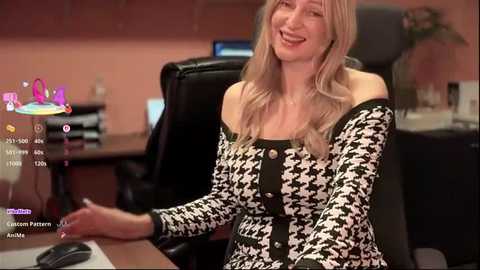 Video of a smiling woman with long blonde hair, wearing a black-and-white houndstooth dress, seated at a desk in an office. Background includes a computer screen and potted plant.