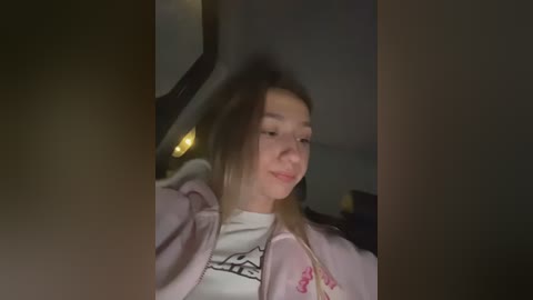 Video of a young Caucasian woman with light skin, long blonde hair, and a white T-shirt with a black graphic, smiling in a dimly lit room.