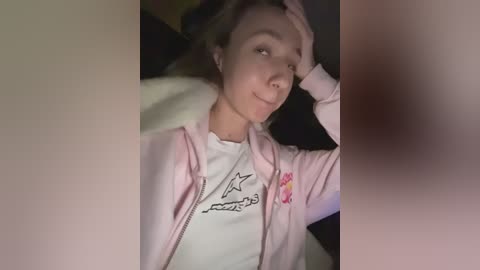A video of a young woman with light skin and brown hair, wearing a pink zip-up hoodie and a white T-shirt, lying on a white pillow, smiling.