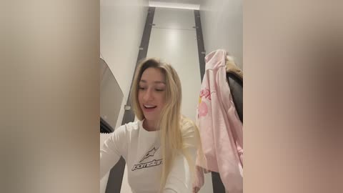A video of a smiling, blonde woman in a white \"Nike\" t-shirt, standing in a narrow, dimly-lit hallway with a pink jacket hanging on the wall.