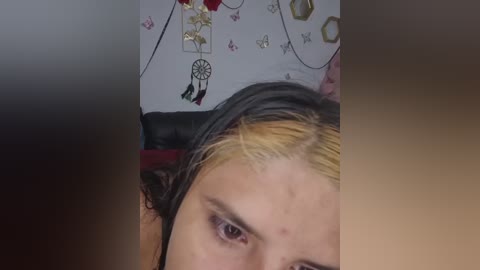 A close-up video of a person with a dyed hairdo featuring blonde highlights and dark roots, lying on a bed with a dreamcatcher wall decoration and a white duvet.