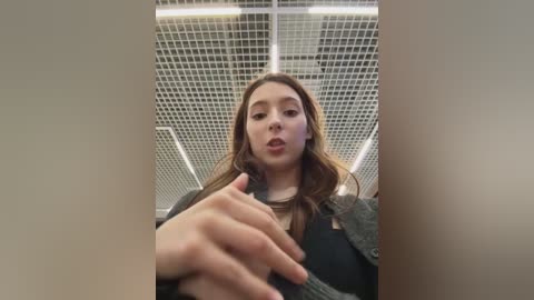 Video of a young woman with light skin and long brown hair, wearing a grey jacket, standing in a modern, grid-patterned ceiling tiled room. She has a neutral expression and is reaching out with her right hand.