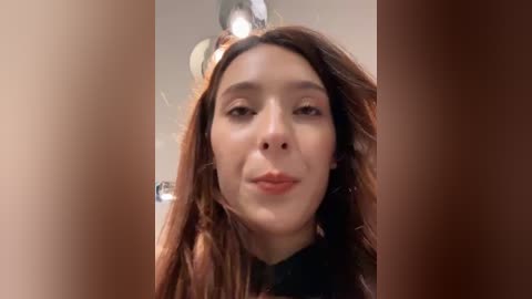 A video of a young woman with long brown hair, light skin, and a neutral expression, taken from a close-up angle, with blurred background.