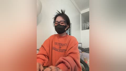 Video of an Asian woman with dark hair, wearing black-rimmed glasses and a face mask, in an orange University of Massachusetts sweatshirt. Background shows a messy bedroom with a bed and bookshelf.