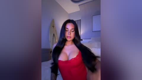 Media: Video of a voluptuous woman with long black hair, wearing a low-cut red lace dress, sitting on a bed in a dimly lit, modern bedroom.