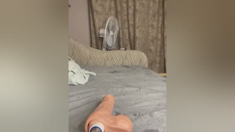 Video of a dimly lit bedroom with a beige bedspread, a white fan on a stand, and a person's hand holding a can of beer in the foreground.