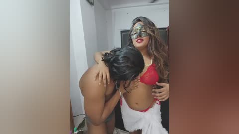 Video: Two women, one in a red lace bra, white skirt, and mask, the other topless, lean toward each other in a dimly lit room with white walls and a door.