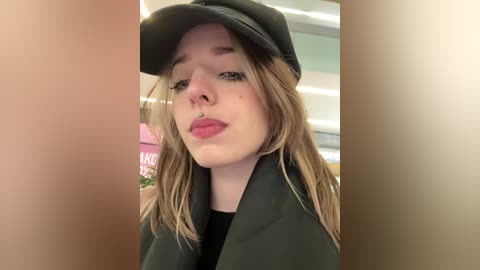 Video of a young woman with pale skin, light brown hair, wearing a black cap and dark jacket, with a hint of pink lipstick, taken indoors with a blurred background.