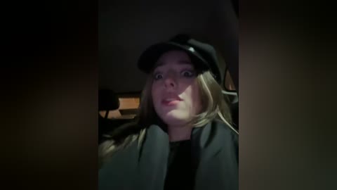 A dimly-lit video of a blonde woman in a black hat and coat, with a neutral expression, sitting in a dimly-lit car interior.