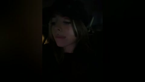 A dimly lit video of a young woman with light skin, wearing a black fur hat, smiling softly. Her hair is long and brown, partially visible. The background is dark, providing a shadowy, intimate setting.