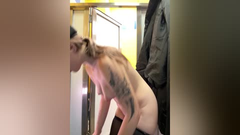 A blurry video of a nude woman with short brown hair, tattoos on her arms, bending over in a dimly lit, narrow hallway.