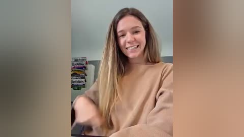 Video of a smiling Caucasian woman with long, straight, blonde hair, wearing a beige sweater, in a brightly lit room with a shelf of colorful boxes in the background.