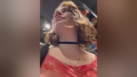 Video of a young woman with curly, shoulder-length brown hair, wearing a black choker and a red floral off-shoulder top, captured from a low angle, indoors with a blurred background.
