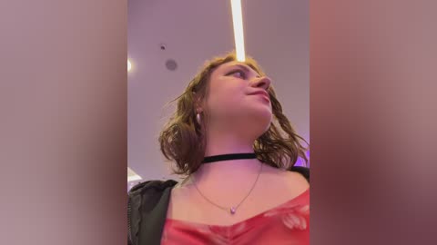 A video of a young woman with shoulder-length, curly brown hair, wearing a black choker and a pink, off-the-shoulder top, gazing upward in a modern, brightly-lit indoor setting.