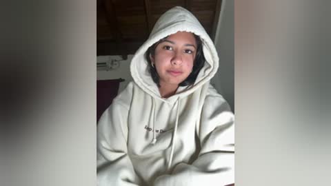 Video of a young Asian woman with light skin, wearing a cream-colored hoodie, smiling gently. Background shows a wooden ceiling and a white wall.