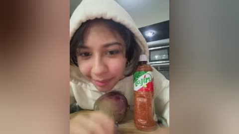Video of a young woman with light brown skin and dark hair, smiling, holding a baby in a white onesie, in a modern kitchen with a red Gatorade bottle and a microwave.