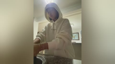 A video of a person wearing a white hoodie, standing in a modern kitchen with beige walls and granite countertops, holding a red object. The person's face is obscured by the hoodie's hood.