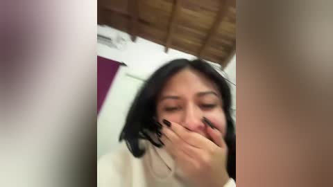 Video of a young Asian woman with shoulder-length black hair, fair skin, and dark nails, covering her mouth with her hand in a bathroom with a wooden ceiling and white walls.