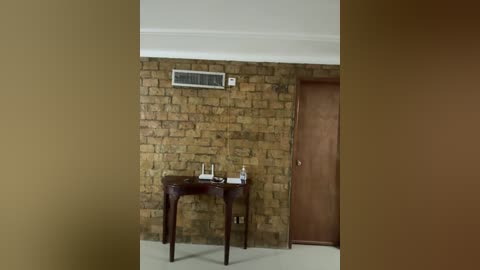 Video of a minimalist room with a brown wooden door, beige walls, and a small wooden table with a white water bottle and a book. The textured stone wall and an air conditioning unit are visible in the background.