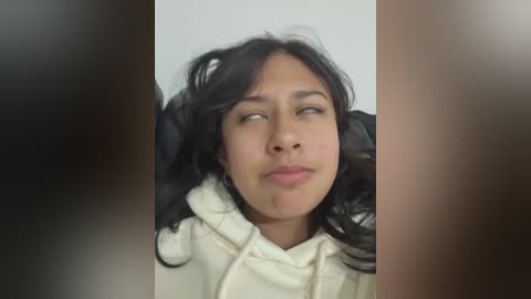 Video of a young woman with light brown skin, dark wavy hair, and light eyes, wearing a white hoodie, looking contemplative against a plain white background.