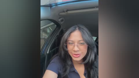 Video of a smiling, light-skinned woman with long black hair, wearing glasses and a black top, seated in the driver's seat of a car.