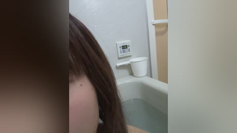 Video of a partially submerged, fair-skinned woman with long brown hair, in a white bathtub, with a white wall-mounted thermostat and a small white trash bin nearby.