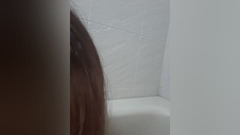 A close-up video of a woman's long, dark brown hair partially obscuring her face, taken in a bathroom with white tiled walls and a white bathtub. The image is slightly blurry and out of focus.