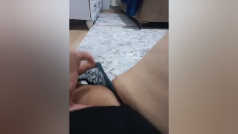 Video of a person's arm in a black shirt, holding a green and white-patterned bra, lying on a light-colored rug in a room with wooden floors and a white dresser.