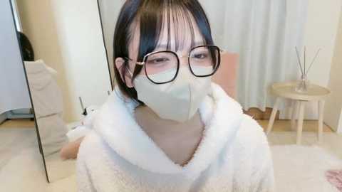 Media: Video of an East Asian woman with straight black hair, wearing glasses, a white mask, and a fluffy white bathrobe, seated in a modern, light-toned bathroom with a mirror, wooden stool, and a white curtain.