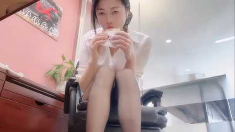 Video of a young Asian woman with light skin and long black hair, wearing a white dress, eating a sandwich while sitting on a black office chair. Background includes a pink wall, a wooden desk, and a glass table.