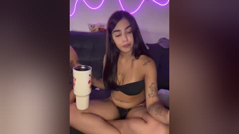 Video of a Latina woman with long black hair, medium breasts, wearing a black bra and panties, holding a white coffee mug, sitting on a dark couch with purple neon lights in the background.