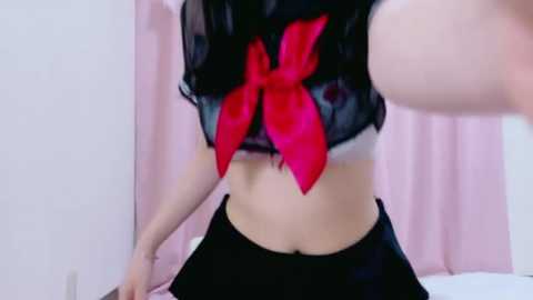 A video of a young woman with pale skin and long black hair, wearing a black, semi-transparent top with a large red bow, and black skirt, in a softly lit room with pink curtains.