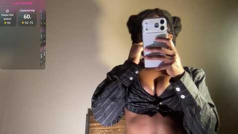A video of a woman with medium skin tone and dark hair styled in buns, wearing an unbuttoned black pinstripe shirt, revealing cleavage. She's taking a selfie with a silver iPhone, positioned indoors against a beige wall.
