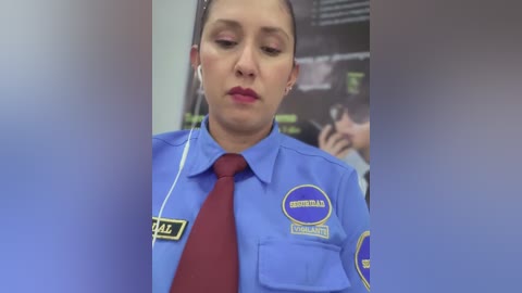 Video of a female airport security officer in a blue uniform with a red tie, name badge, and yellow ID patch, wearing white earbuds, looking down, in a blurred airport setting.