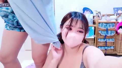 Video of a young East Asian woman in a gray bra, taking a selfie in a bedroom. She has straight black hair, light skin, and a slender build. The background shows a messy wooden dresser with clothes.