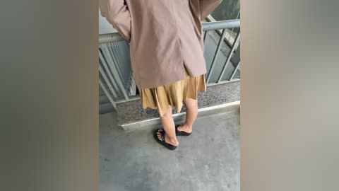 A video of a person with light skin, wearing a beige blazer, mustard-yellow skirt, and black flip-flops, standing on a gray concrete floor near a metal railing.