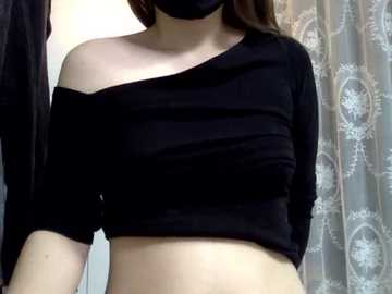 Video of a woman with light skin wearing a black off-shoulder crop top and black face mask, standing in a bathroom with a floral-patterned shower curtain.