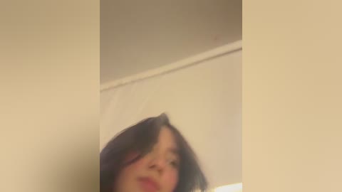 A video of a person with shoulder-length dark hair, standing in a dimly lit room with beige walls. The image is slightly blurred, making details hard to discern.