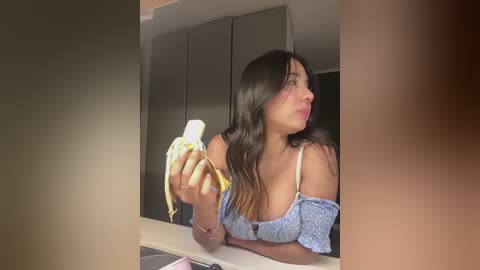 Video of a young woman with long, straight black hair and a light skin tone, wearing a blue off-shoulder top, holding a peeled banana in her left hand. She gazes to the right, with a neutral expression.