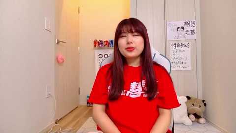 Video of a smiling, fair-skinned woman with long, straight brown hair wearing a red t-shirt with Japanese characters. She sits on a white chair in a minimalist room with a wooden floor and a plush bear toy beside her.