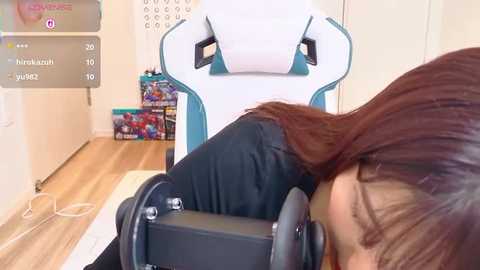 Video of a young person with shoulder-length brown hair, wearing black clothing, sleeping on a gaming chair with a white and teal design. Background features a wooden floor and a wall with a calendar and a box of games.