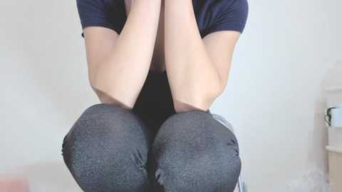 Video of a person, likely female, kneeling with hands clasped, wearing dark grey leggings and a navy t-shirt, against a plain white wall, with a small potted plant in the background.