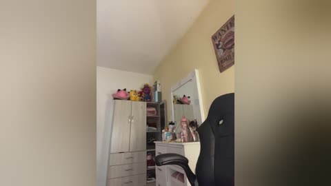 Video of a cozy bedroom with light yellow walls, a black gaming chair, a mirrored vanity table, and a dresser filled with pink and purple toiletries, against a white background.