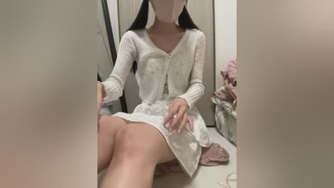 Video of an Asian woman with long black hair, wearing a white, knitted cardigan and skirt, sitting on a white floor in a dimly lit room, holding a pink towel.